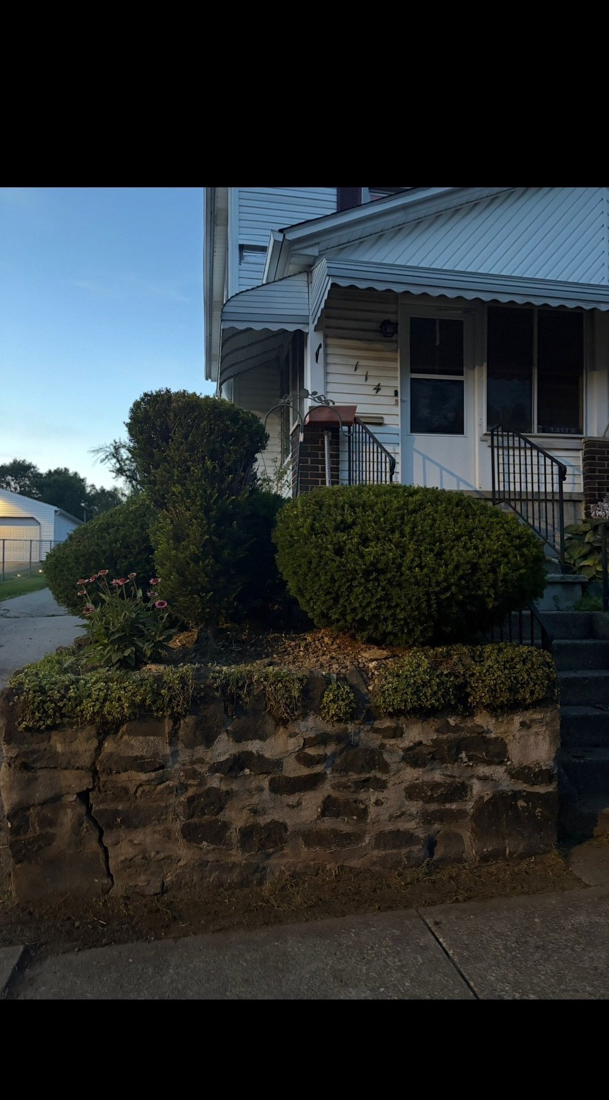 Front of residential house with stone retaining wall, green shrubs, and metal roof on clear day