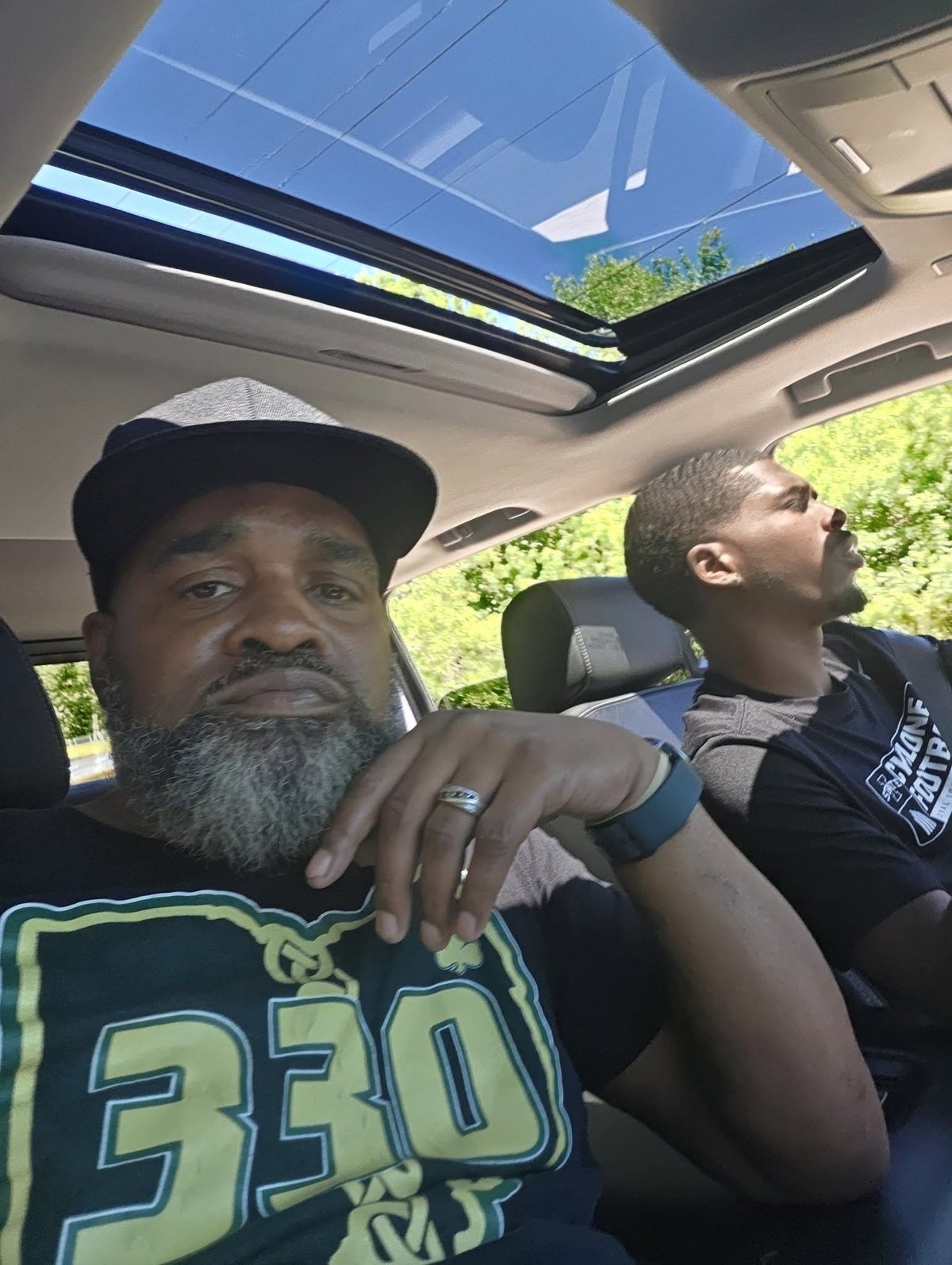 Two people sitting in a car with open sunroof, man on left wearing black cap and graphic shirt